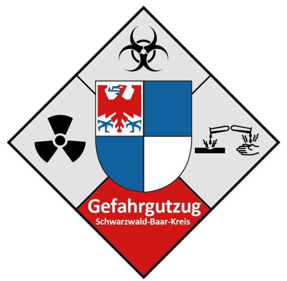 Raute mit mittigem blau-weiß gevierteiltem Wappenschild (roter Adler oben links), umgeben von Biohazard-, Radioaktivitäts- und ätzend-Symbolen; rotes Feld unten mit weißer Schrift "Gefahrgutzug Schwarzwald-Baar-Kreis".