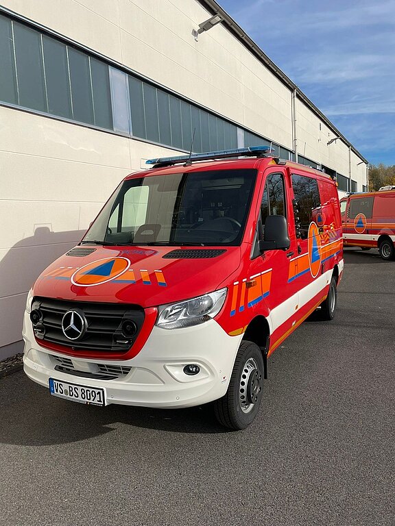 Rotes-weißes Mercedes-Einsatzfahrzeug des Katastrophenschutzes mit blau-orangeem Logo, geparkt vor einer Industriehalle.